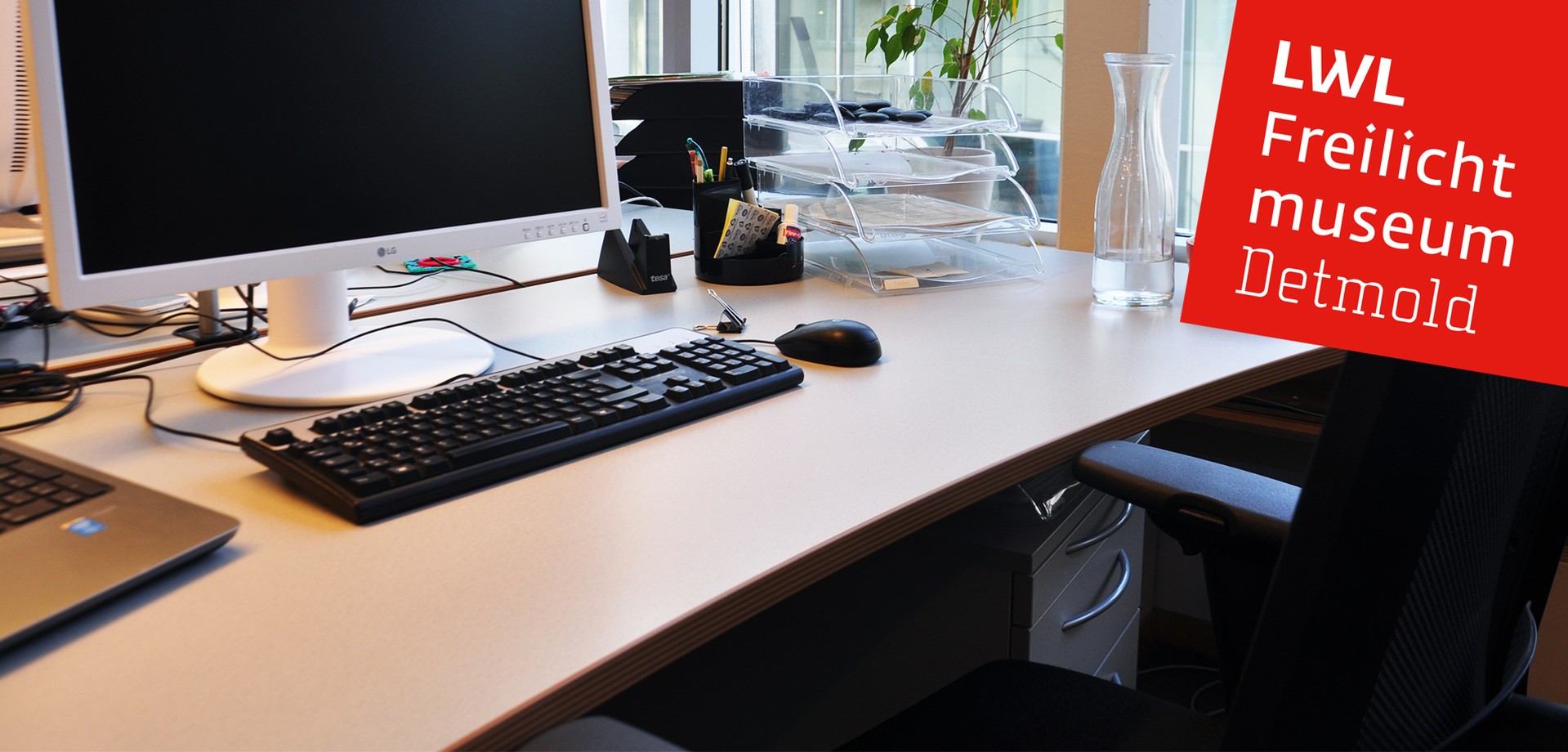 Einen Arbeitsplatz in einem unserer Büros. Auf dem Bild ist ein PC, eine Tastatur, eine Maus und diverses Schreibtischzubehör.