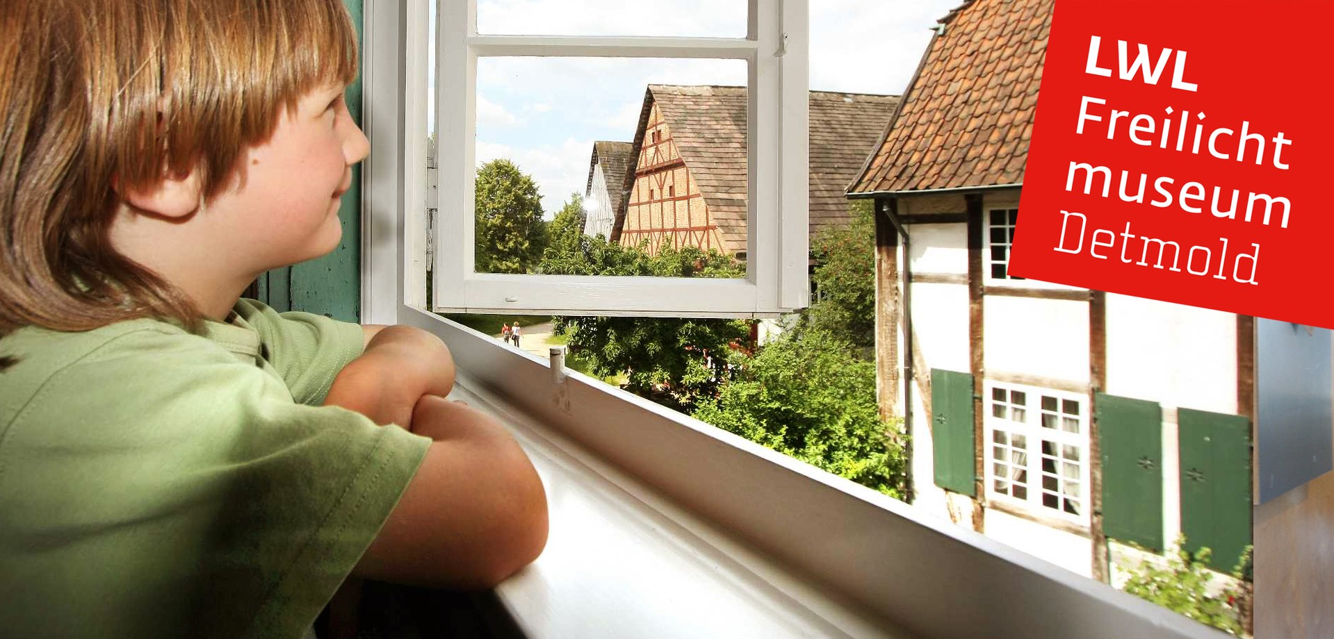 Einen Jungen, der aus einem Fenster schaut. Hinter dem Fenster sind Häuser des Museums.