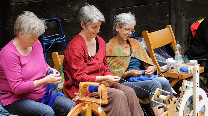 Three women sitting at a spinning wheel. There is a yarn spool on the spinning wheel. They sew something out of it.