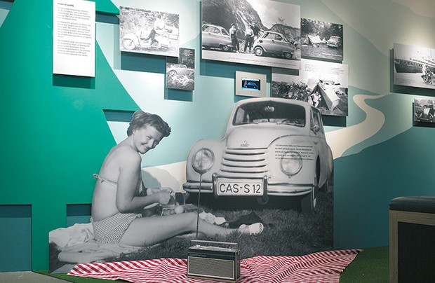 Eine Ausstellungswand mit Bildern historischer Autos und Motorräder sowie eine Frau im Bikini, die auf einer Wiese vor ihrem Auto sitzt.