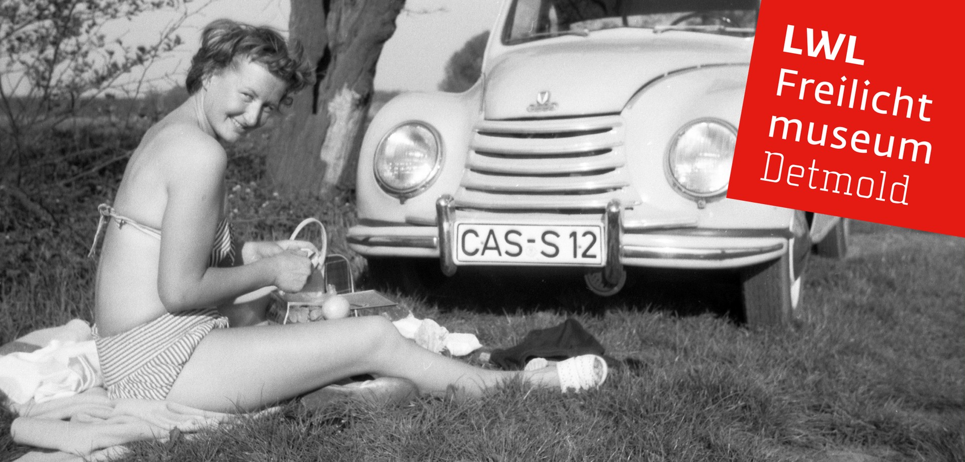 Eine Frau im Bikini, die auf einer Wiese sitzt. Neben ihr ein historisches Auto
