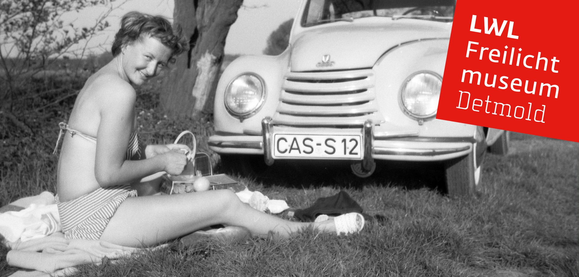 Eine Frau im Bikini, die auf einer Wiese sitzt. Neben ihr ein historisches Auto
