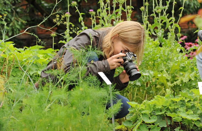 Eine Frau hockt in einem Garten zwischen Pflanzen und macht ein Foto von einer Pflanze.