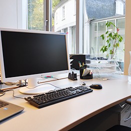 Einen Arbeitsplatz in einem unserer Büros. Auf dem Bild ist ein PC, eine Tastatur, eine Maus und diverses Schreibtischzubehör.