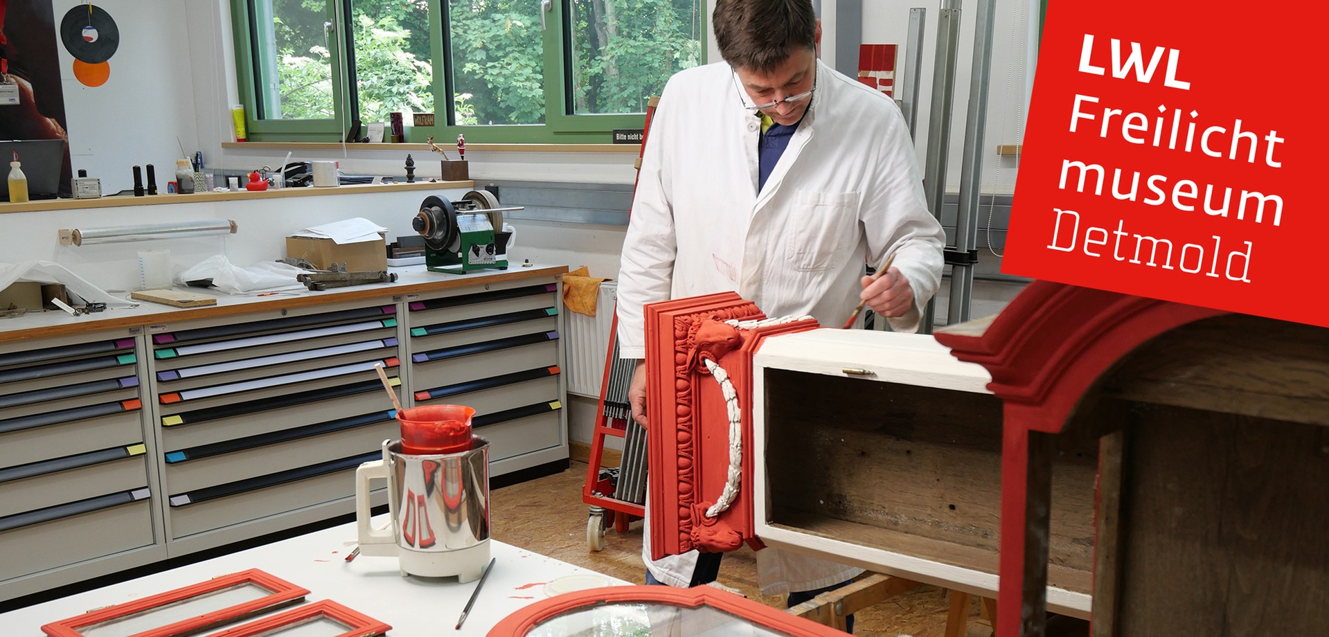 Ein Restaurator mit braunem Haar und Lesebrille steht an einem Tisch und trägt mit einem Pinsel Farbe auf eine rote Standuhr auf.