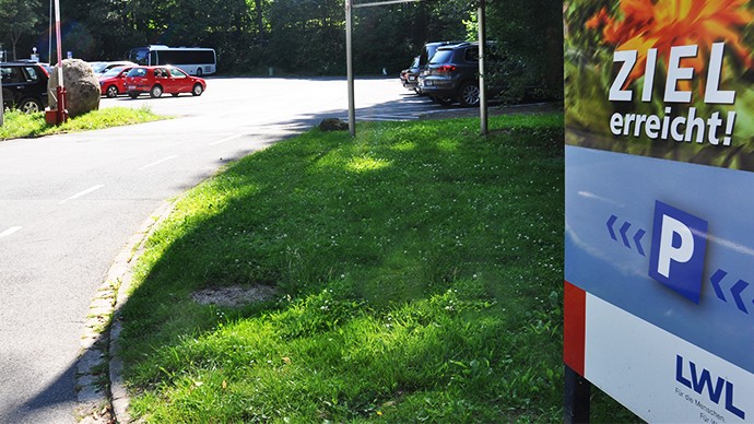 Die Einfahrt zum Parkplatz. Rechts im Bild das Parkplatzschild mit der Aufschrift "Ziel erreicht". Auf dem Parkplatz stehen einige Autos.