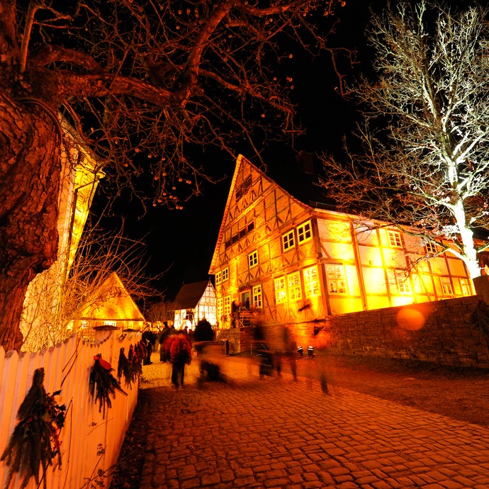 An einem Weg aus Kopfsteinpflaster stehen mehrere Fachwerkhäuser, die gelb und weiß beleuchtet sind. An einem Zaun links hängen Tannenbündel. (Enlarged picture view opens)