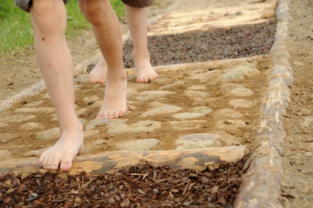 Zwei Kinder laufen über einen Barfußpfad, der aus verschiedenen Materialien besteht: Stein, Holz, Rindenmulch, Kies. Zwei Kinder laufen über einen Barfußpfad, der aus verschiedenen Materialien besteht: Stein, Holz, Rindenmulch, Kies.