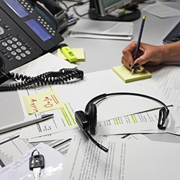 Auf einem Schreibtisch liegen Papiere und ein Kopfhörer. Im Hintergrund ist ein Telefon zu sehen und rechts eine Hand, die etwas auf einen Block notie
