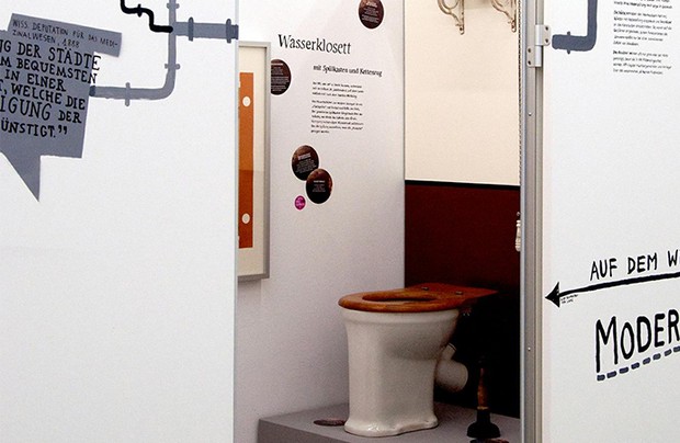 Eine Toilette. Rundherum Wände, auf denen Informationen zur Ausstellung stehen und Rohre aufgemalt sind.