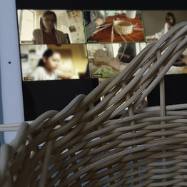 Vor dem Monitor eines Tablets, auf dem gerade Teilnehmende eines Flechtkurses zu sehen sind, steht ein halbfertiges geflochtenes Körbchen.