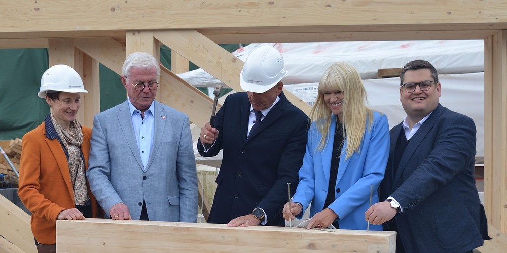 Fünf Personen stehen hinter einem Holzblock; eine Person schlägt einen Nagel ein.